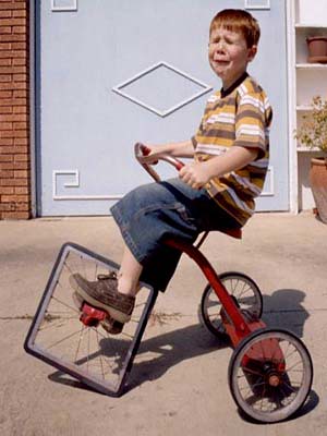 Boy riding tricycle with square front wheel. Boy is not happy